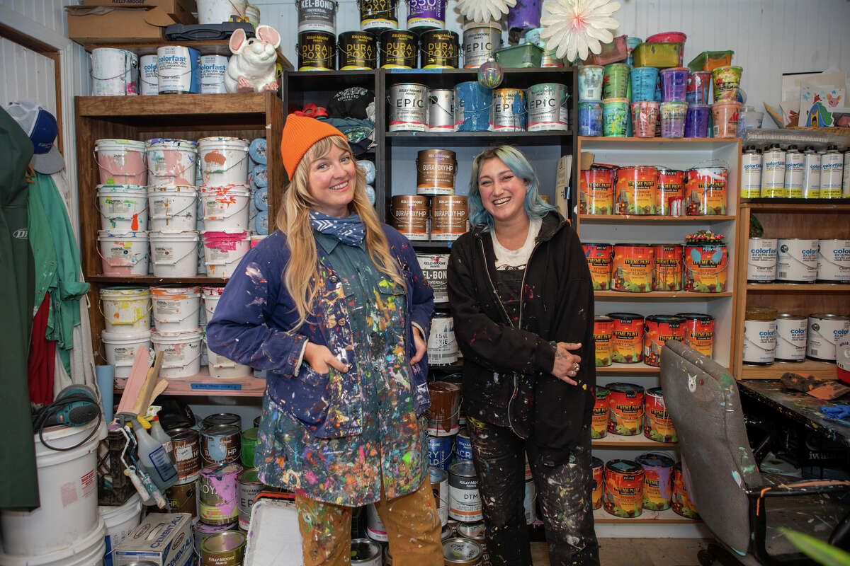 La directora de arte y restauración de Children's Fairyland, Shannon Taylor, a la izquierda, y el pintor Ari Bird se paran frente a las latas de pintura que usan en el taller del departamento de arte en Oakland, California, el 8 de junio de 2022. Trabajan juntos para restaurar y mantener el tema atracciones del parque en plena forma.