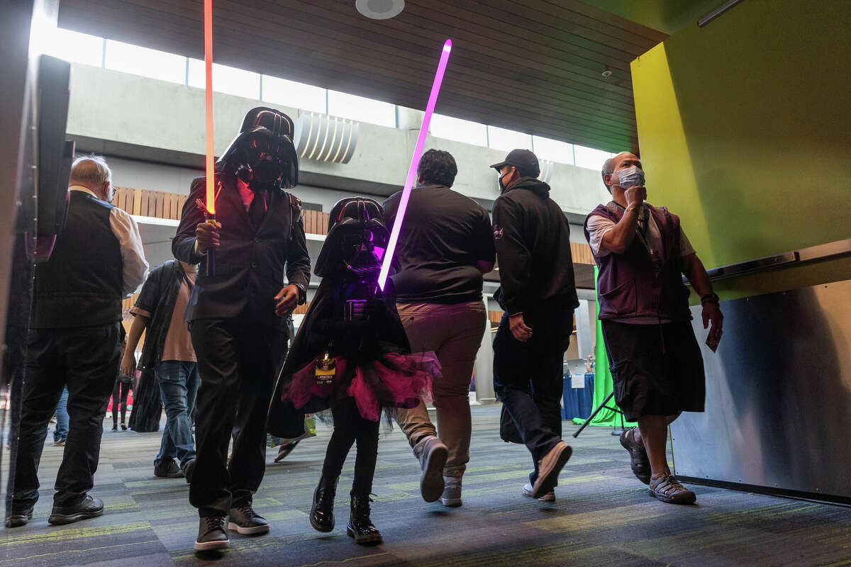 Ernie Campos y su hija Elianna Campos, ambos con máscaras de Darth Vader, caminan por SiliCon con Adam Savage blandiendo sables de luz en el Centro de Convenciones McEnery de San José el 27 de agosto de 2022. 