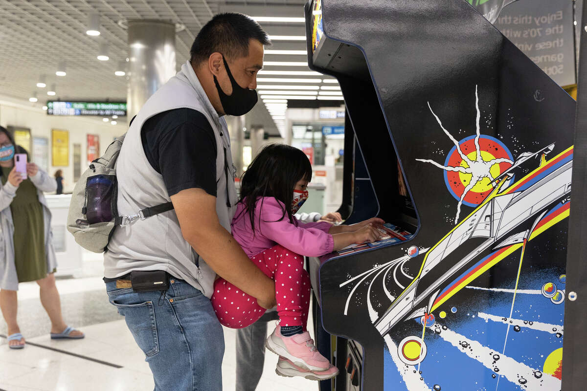 Matt Lee sostiene a su hija frente a una máquina recreativa antigua para que pueda jugar Asteroids en la estación BART de Powell Street en San Francisco el 3 de septiembre de 2022. 