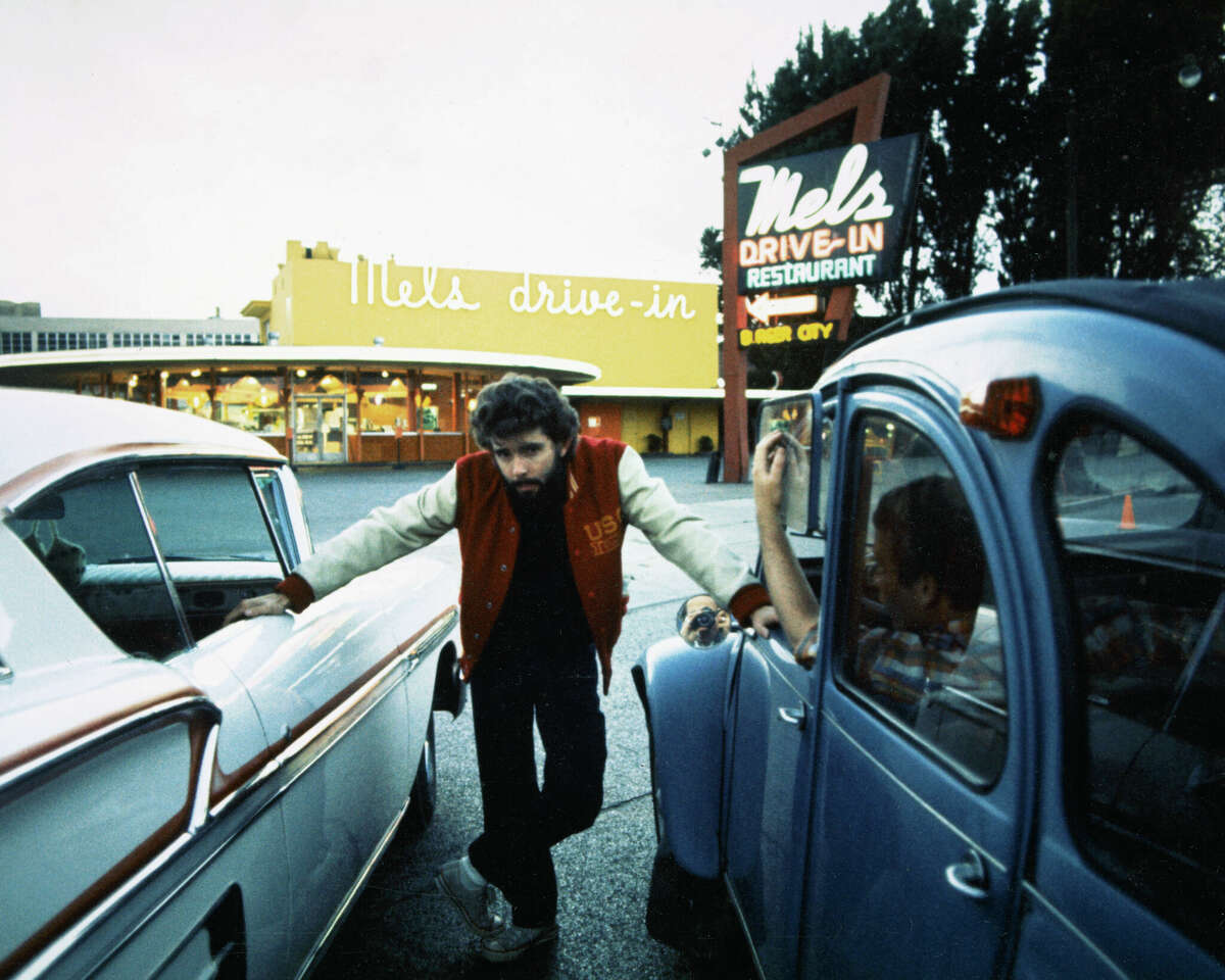 George Lucas, director de cine estadounidense, posando entre dos coches, con "Autocine de Mel" en el fondo, en el set de "grafiti americano," San Francisco, 1973. 