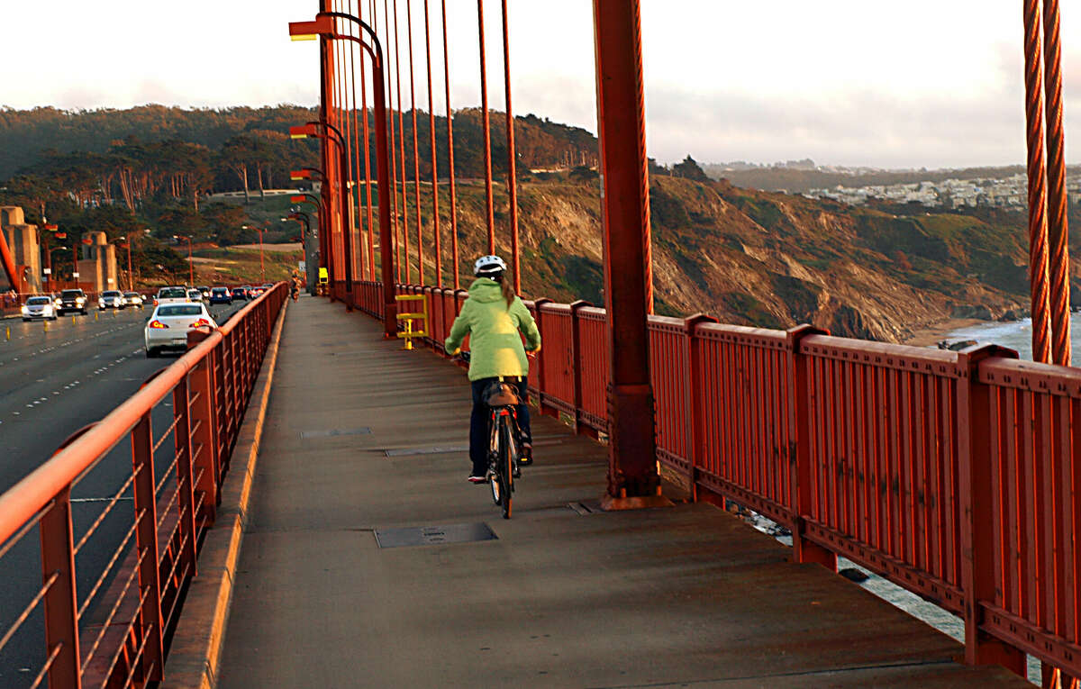 Una mujer cruza en bicicleta el puente Golden Gate de San Francisco al atardecer, mientras el intenso tráfico de automóviles cruza el puente. Otros dos puentes a lo largo del sendero de la bahía de San Francisco no tienen senderos para bicicletas o peatones en la actualidad. 